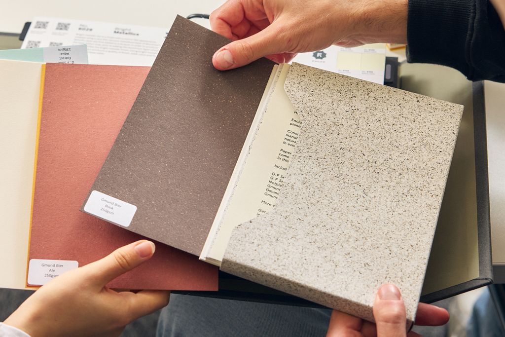 Hands holding a dark brown flecked paper sample and a black and white flecked paper sample